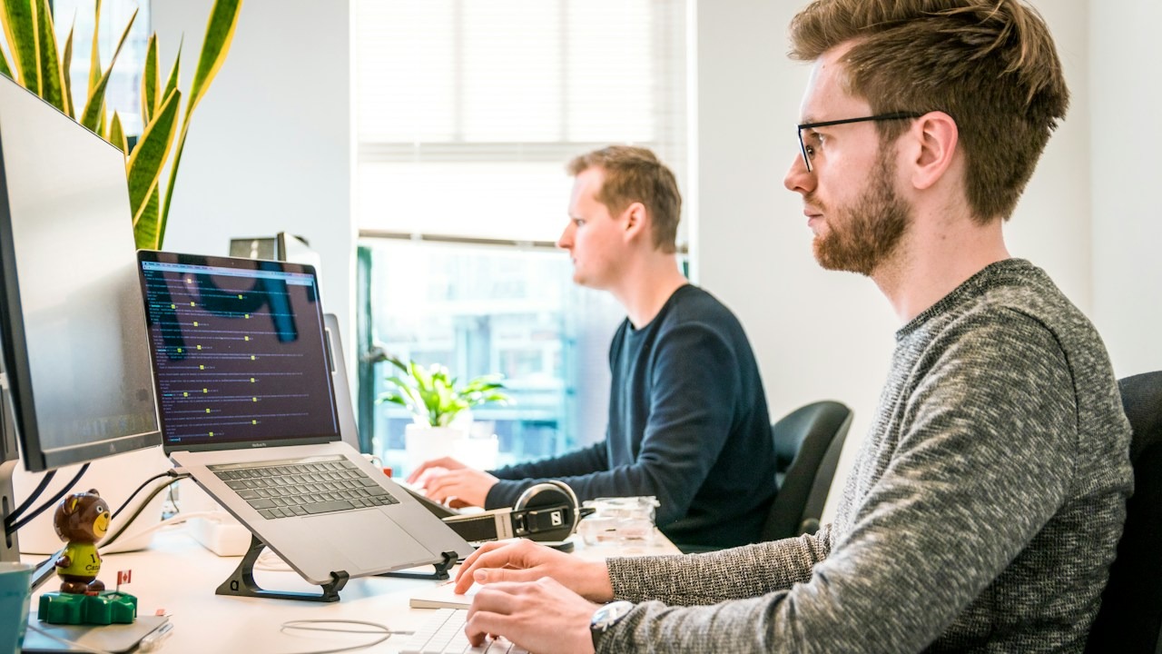 Engineer reviewing code on a laptop in a bright modern office while a teammate works nearby.